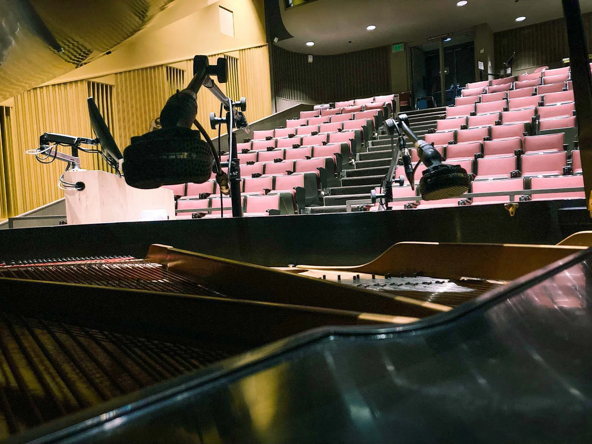 Microphones over an open grand piano in an empty recital hall with pink seats.